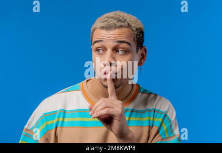 Handsome man with gesture of shhh, secret, silence, conspiracy, gossip concept. Smiling guy holding finger on lips, blue studio background.  Stock Photo