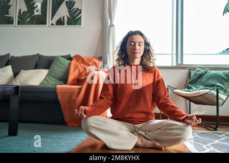 Young calm woman wearing headphones doing yoga meditation at home. Stock Photo