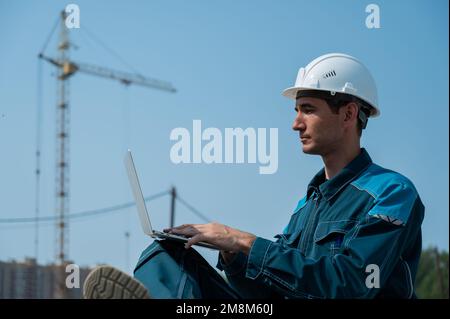 Caucasian male builder in hardhat sits on floor slabs and uses laptop at construction site.  Stock Photo