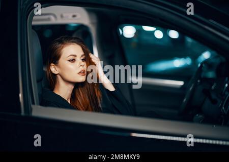 a stylish, luxurious woman sits in a black car at night, touching her face with a thoughtful expression. Close horizontal photo Stock Photo