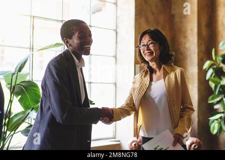 Two diverse women one African American with short hair in casual attire shake hands and the other Asian laugh in an office with natural light coming f Stock Photo