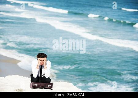 Lost in a sea of worries. Young businessman sitting on a cliff above the ocean with his hands in his hair. Stock Photo