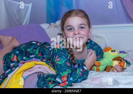 Smiling six year old girl missing front teeth lying on her bed with her stuffed animals. Stock Photo