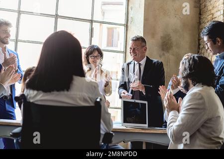 A diverse group of people, standing and sitting around a table applauding a man holding a model of a wind turbine, in an office. Celebrating teamwork Stock Photo