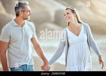 Happy and loving mature caucasian couple enjoying a romantic walk at the beach together on a sunny day. Cheerful affectionate husband and wife holding Stock Photo