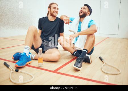 Two young athletic squash players taking break after playing court game. Full length of laughing mixed race and caucasian athlete sitting together Stock Photo