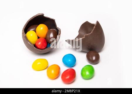 Cracked chocolate easter egg full of colorful small round candies and an empty half of the egg on a white background with colorful candies. Chocolate Stock Photo