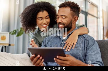 Mixed race couple smiling while using a digital tablet together at home. Cheerful hispanic boyfriend and girlfriend relaxing and using social media on Stock Photo