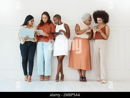 Group of five diverse young businesswomen standing against a wall in an office and using tech. Happy colleagues talking and using technology while Stock Photo