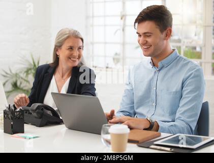 Mature caucasian businesswoman talking to a young male colleague while working on a laptop together at work. Two colleagues laughing while sitting at Stock Photo