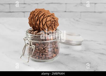 Pine nuts in a jar and a pine cone in the kitchen on a light background. The concept of organic food and protein food Stock Photo