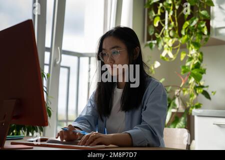 Focused Asian woman IT developer wearing glasses writing code on computer while working from home Stock Photo