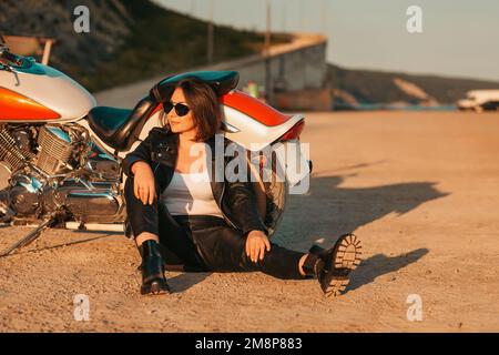 Pretty confident woman in a leather motorcycle outfit sitting on the ground near a motorcycle. The concept of the Motorcyclist Day and feminism. Stock Photo