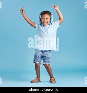 Full length of a funny little boy dancing while listening to music with wireless headphones and wearing casual clothes against a blue studio Stock Photo