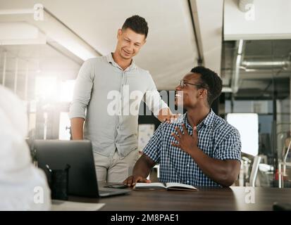 Two diverse businessmen talking at a desk discussing new project on laptop. Mentor complimenting colleague on a job well done and being a fast learner Stock Photo