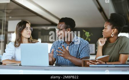 Three young happy businesspeople having a meeting while sitting at a table and working on a laptop at work. Business professionals talking and Stock Photo