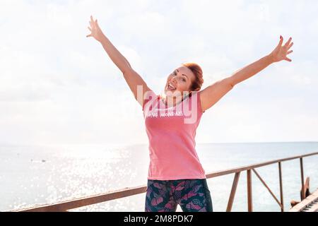 An adult woman in sportswear smiles happily and poses with her hands raised in the air against the sky and sea. Copy space. The concept of sport and a Stock Photo