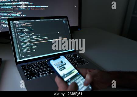Programmer checking phone while writing code on laptop Stock Photo
