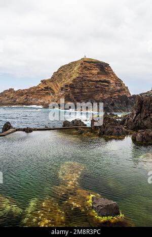 Atlantic Ocean with natural pool and rocky isle with lighthouse near Porto Moniz in Madeira during cloudy springtime day Stock Photo