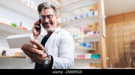 Drug store pharmacist holding a box of pills and talking to a patient on the phone. Senior healthcare worker reading medication label to confirm a pre Stock Photo