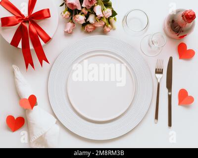 Valentines Day tablesettings concept on white background. Empty white plates, gold cutlery, rose wine bottle, glasses, flowers bouqet, gift box. Copy space. Topview flatlay Stock Photo