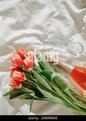 Bunch of red tulips, rose wine and champagne glasses on white bed sheets. Valentines day, date, march 8 concept. Spring flowers and bottle. Top view or flat lay. Copy space. Vertical Stock Photo