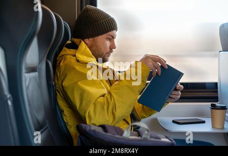 Bearded man in winter yellow jacket and hat reading book while traveling by train sitting inside train. Stock Photo