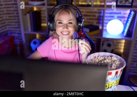 Young blonde woman streamer playing video game eating popcorn at gaming room Stock Photo