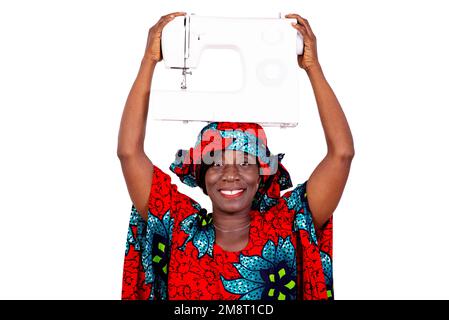 a seamstress in traditional dress standing against white background carrying a sewing machine on her head and looking at the camera smiling. Stock Photo