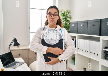 Young hispanic woman working at dietitian clinic puffing cheeks with funny face. mouth inflated with air, catching air. Stock Photo