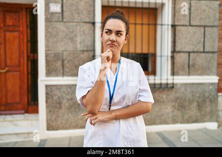 Young hispanic woman wearing doctor id card thinking concentrated about doubt with finger on chin and looking up wondering Stock Photo