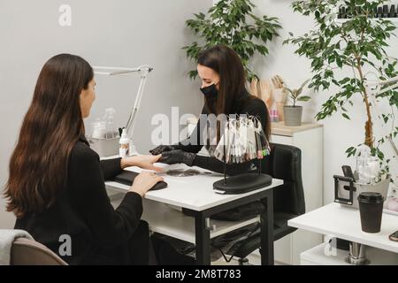 Nail service. Happy customer getting her nails done at beauty salon. Manicure specialist wearing protective mask doing nails for woman client in Stock Photo