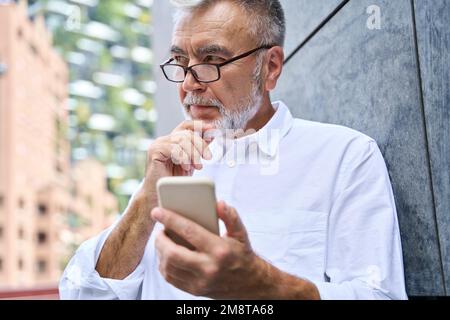 Old senior professional business man holding phone using smartphone thinking. Stock Photo