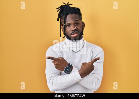 African man with dreadlocks wearing turtleneck sweater over yellow background pointing to both sides with fingers, different direction disagree Stock Photo