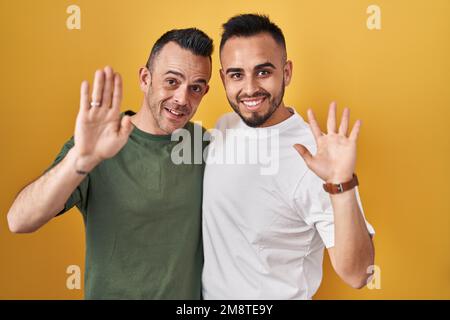 Homosexual couple standing over yellow background waiving saying hello happy and smiling, friendly welcome gesture Stock Photo