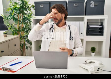 Handsome middle age doctor man working at the clinic smelling something stinky and disgusting, intolerable smell, holding breath with fingers on nose. Stock Photo