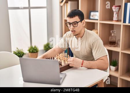 Young arab man playing chess online scared and amazed with open mouth for surprise, disbelief face Stock Photo