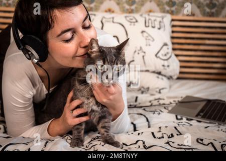 Beautiful woman plays with the cat on the bed. The young woman listens to music on headphones while she is petting her cat. Stock Photo
