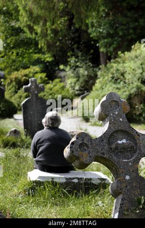 Woman sitting near medieval stone cross of the Glendalough monastery in the Wicklow mountains in Ireland Stock Photo