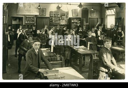 Original and clear early 1900's Edwardian postcard of junior Edwardian schoolboys in woodwork carpentry class tools, The boys are wearing suits with large starched, detachable collars. On the wall are displays of completed projects - plate racks, chess board, picture frame. from the Photographer E. Fenske, 52 Osborne Road, Thornton Heath, South London, U.K. circa 1905 / 1910. Stock Photo