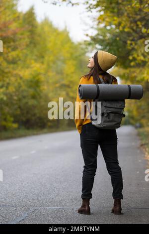 Young brunette woman with backpack stands on autumn road Stock Photo