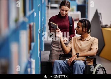 Graphic portrait of smiling young woman assisting student with disability in library and choosing books together Stock Photo