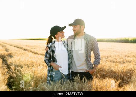 A couple of farmers in plaid shirts and caps stand embracing on agricultural field of wheat at sunset Stock Photo