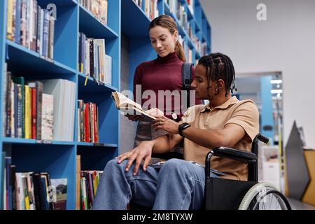 Portrait of black young man using wheelchair in library and choosing books with friend helping Stock Photo