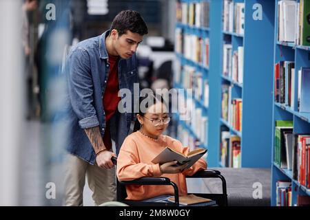 Portrait of Asian girl with disability in library choosing books with friend assisting, inclusivity concept Stock Photo