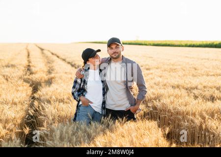 A couple of farmers in plaid shirts and caps stand embracing on agricultural field of wheat at sunset Stock Photo