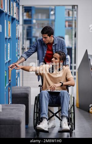 Vertical full length portrait of young man assisting student with disability in library and choosing books together Stock Photo