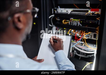 Close up of black man working with server cabinet in data center and taking notes on clipboard, copy space Stock Photo