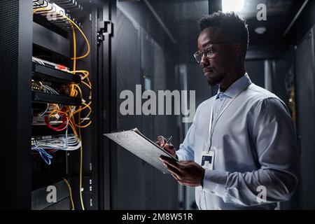 Waist up side view portrait of network engineer working with server cabinet in data center and taking notes on clipboard, copy space Stock Photo