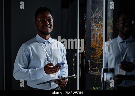 Waist up portrait of smiling black man as network engineer standing by server cabinet in data center and looking at camera, copy space Stock Photo
